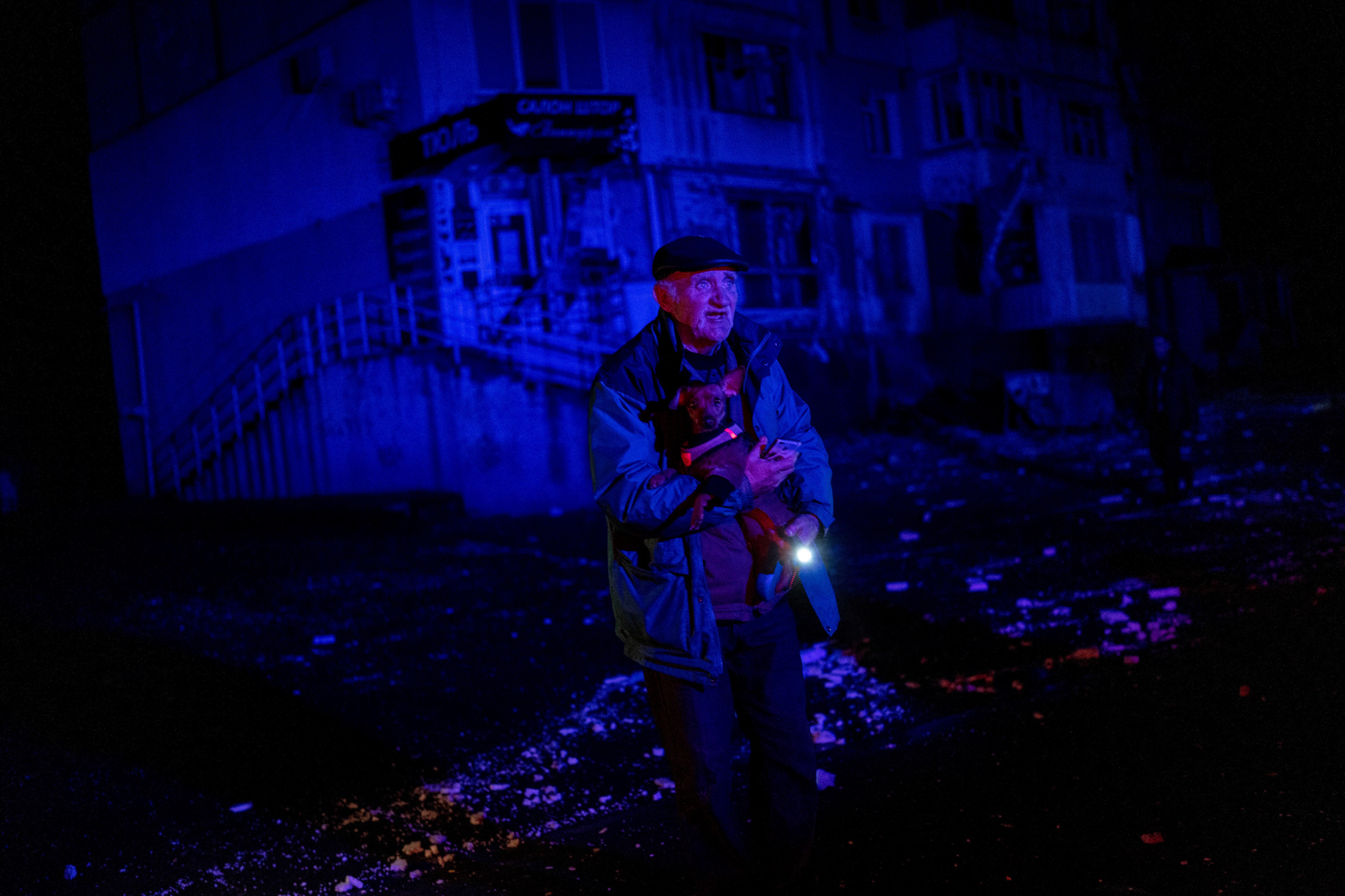 A man stands in the dark holding a torch with rubble around him and damaged buildings behind him. 