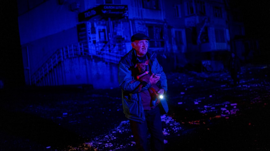 A man stands in the dark holding a torch with rubble around him and damaged buildings behind him. 