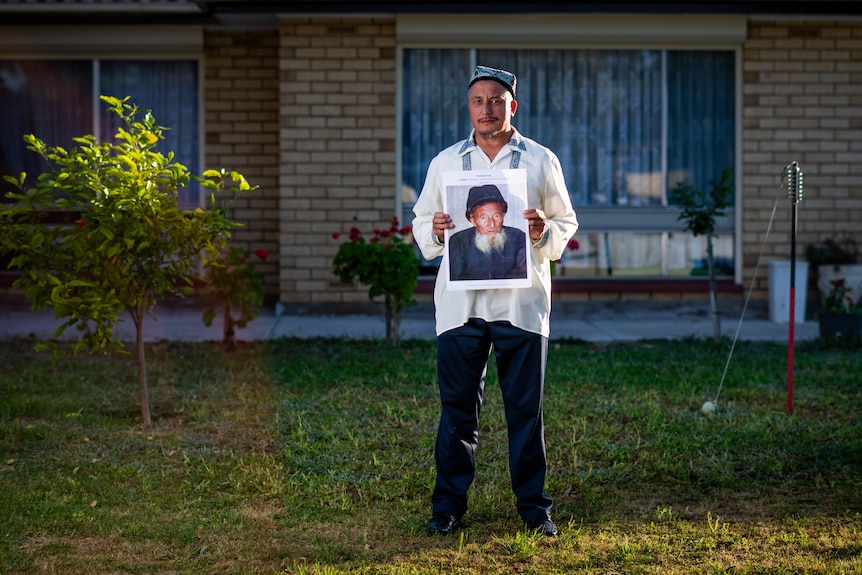 A man on a lawn holds a photo of a an elderly man 
