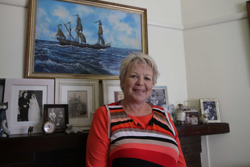 A smiling woman stands in front of a painting of a ship in her loungeroom.