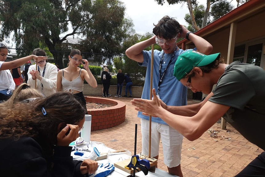 A group of teenagers watch a male trying to make fire by twirling a stick