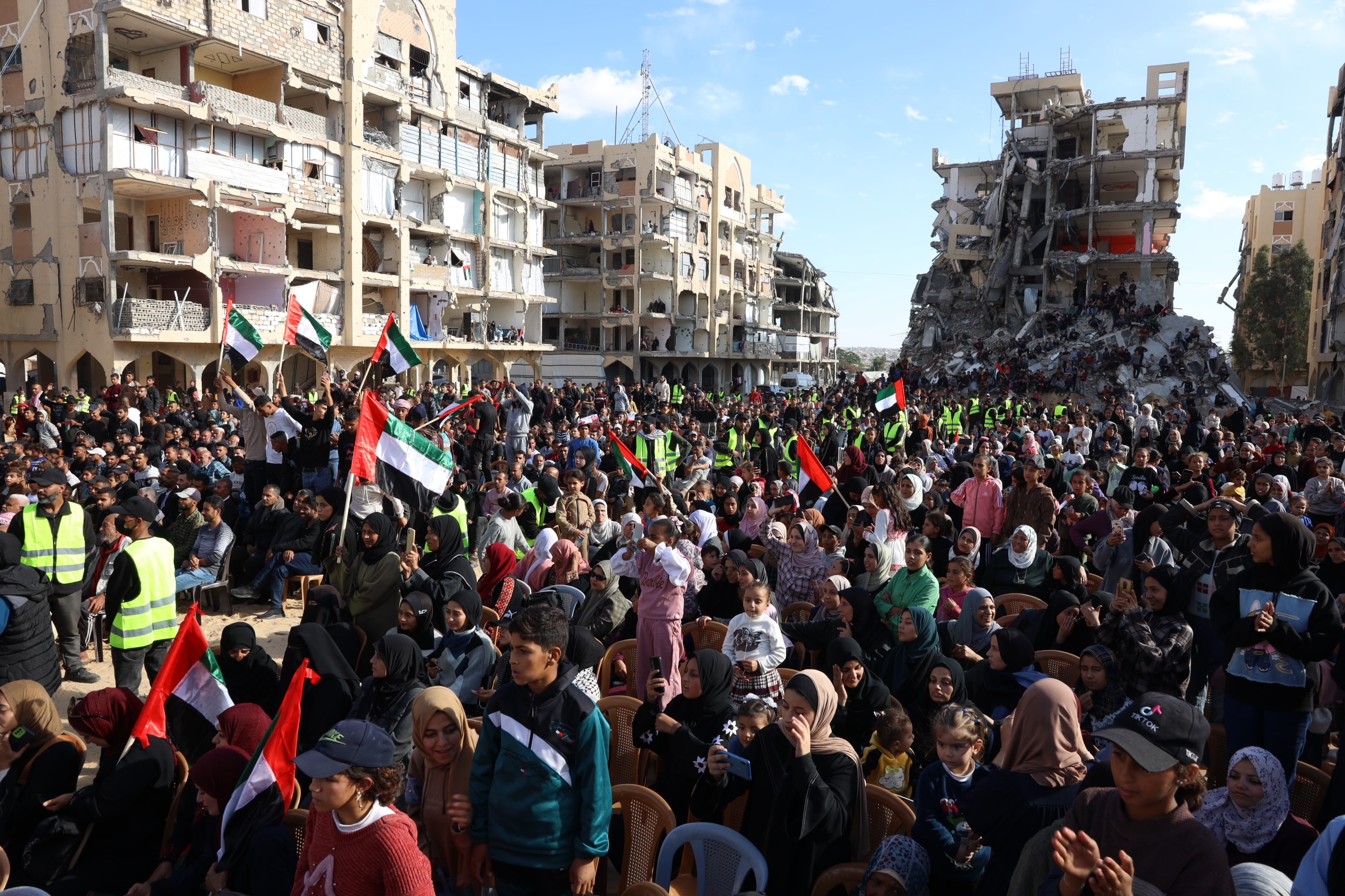 Crowds of people, some sitting, others standing carrying flags and more on top of collapsed buildings behind them.
