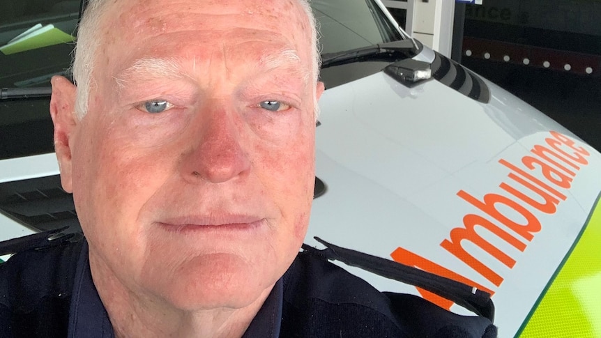 older man in paramedic uniform takes a selfie in front of an ambulance