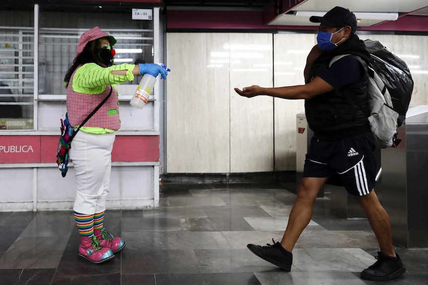 A woman dressed as a clown extends a spray bottle out in front of her to spray a man's extended hand