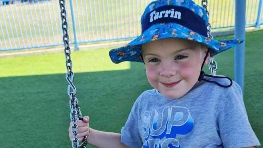 Toddler Tarrin-Macen O’Sullivan smiling and wearing a hat while on a swing.  