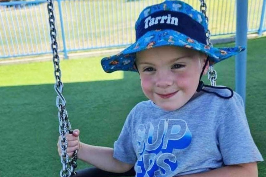 Toddler Tarrin-Macen O’Sullivan smiling and wearing a hat while on a swing.  