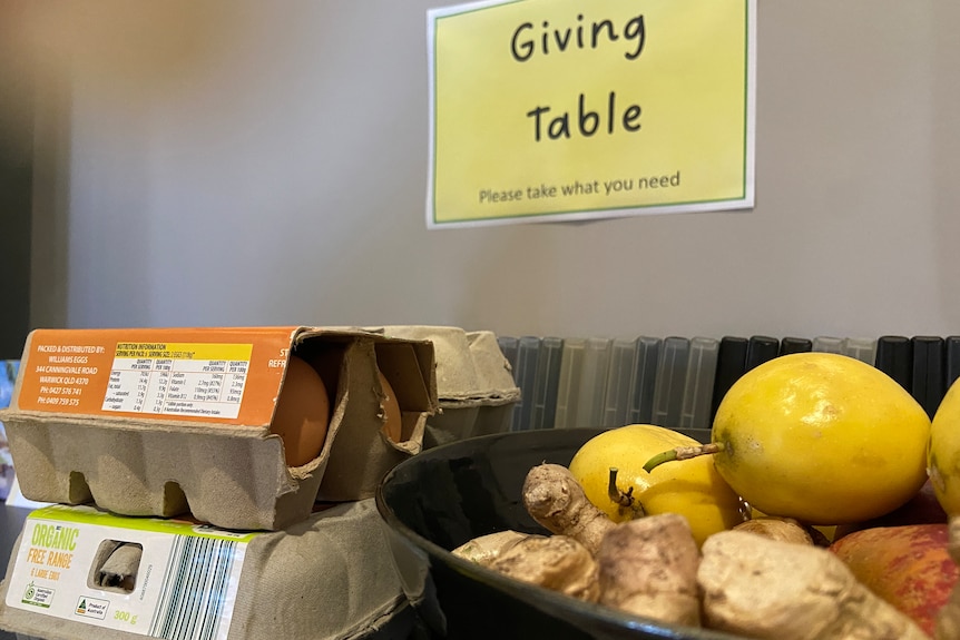 Una pequeña mesa contra una pared.  Un cartel dice Mesa de donaciones.  Sobre la mesa hay medio cartón de huevos y un bol con jengibre y limones.
