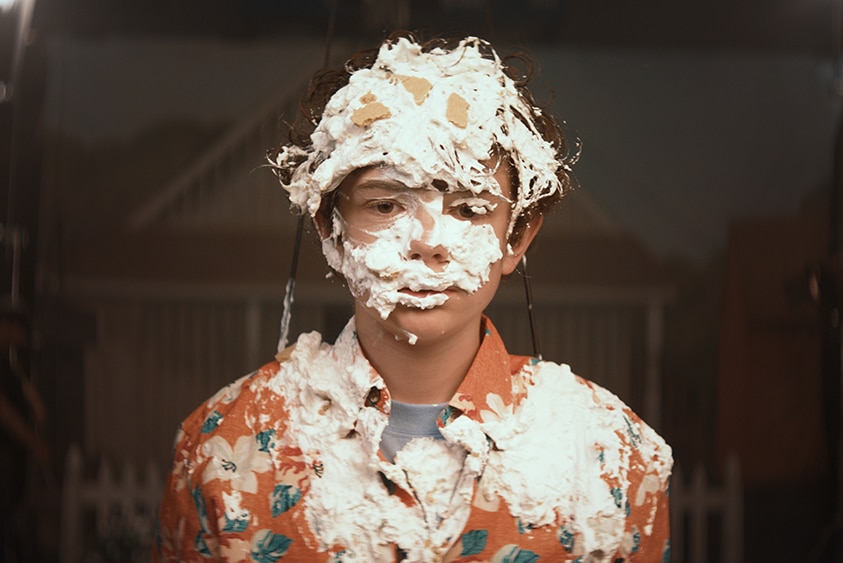 A teenage boy wearing orange hawaiian shirt stands, his torso and head covered in bits of cream and pie.