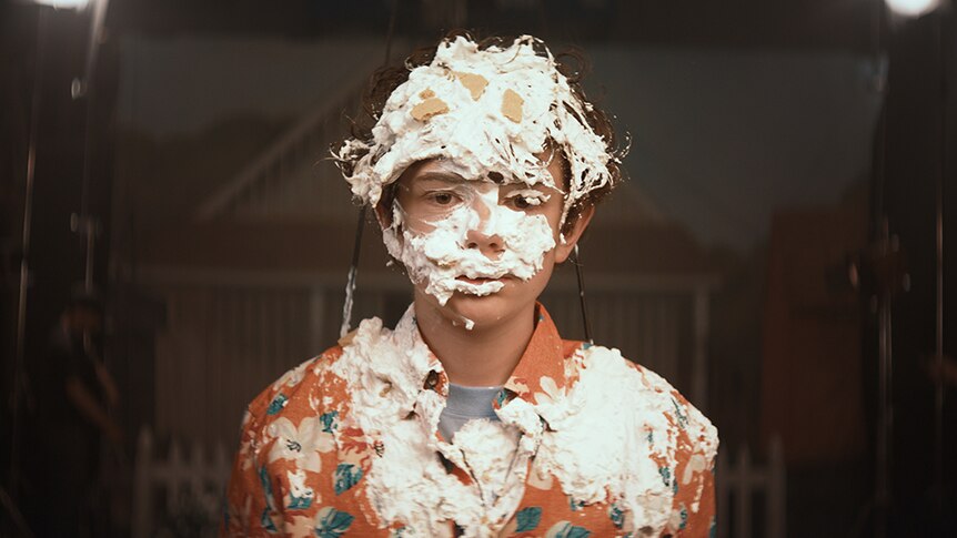 A teenage boy wearing orange hawaiian shirt stands, his torso and head covered in bits of cream and pie.