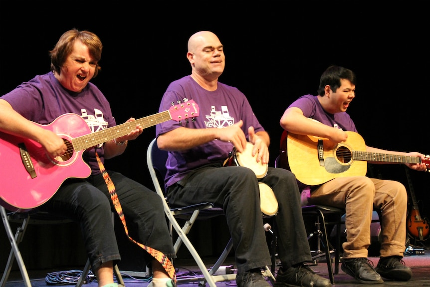 A woman and two men on stage singing and playing instruments.