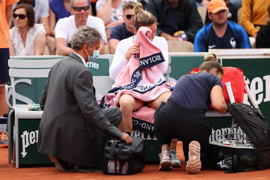 A tennis player sits with a towel to her face as two doctors assess her health on the court
