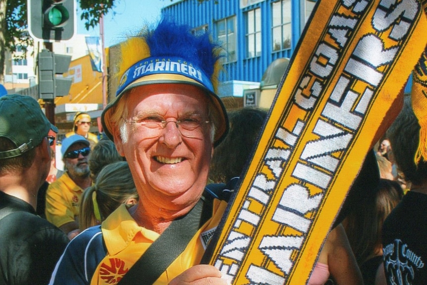 An older man in soccer gear holding an inflatable bat that says "Mariners" on it.