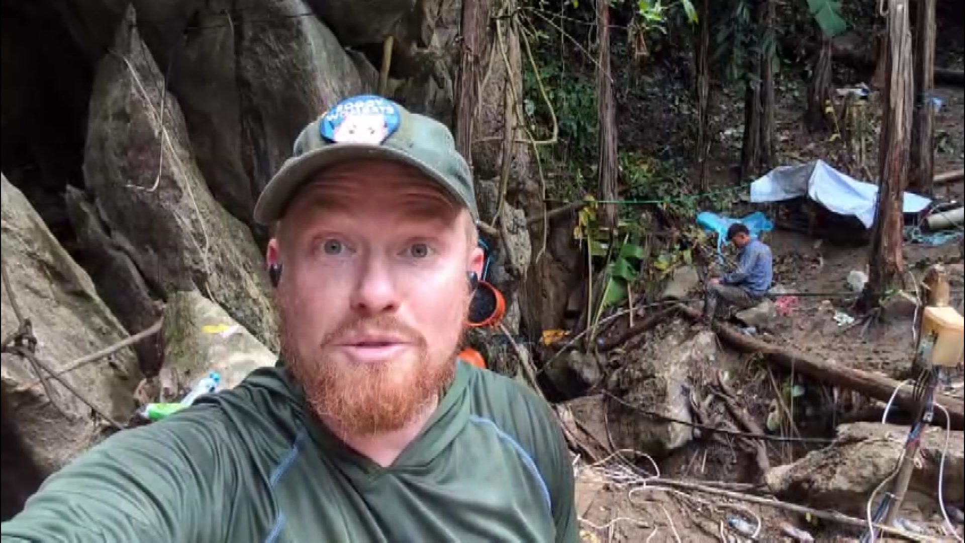 A man wearing a green cap and a green wet suit stands against a jungle backdrop