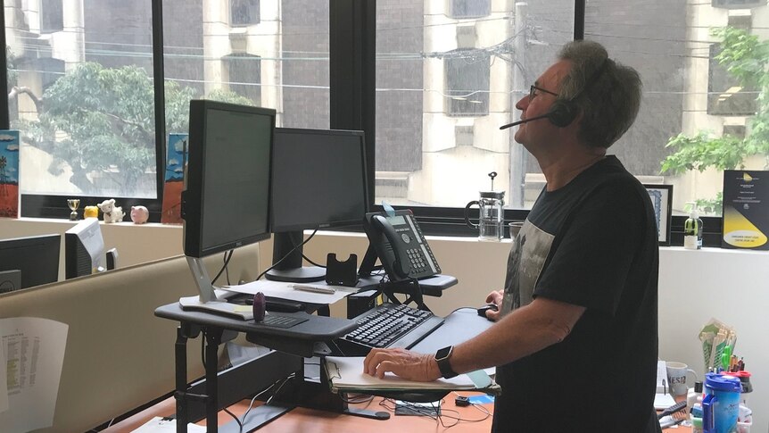 Man standing at raised work desk on the phone at a computer.
