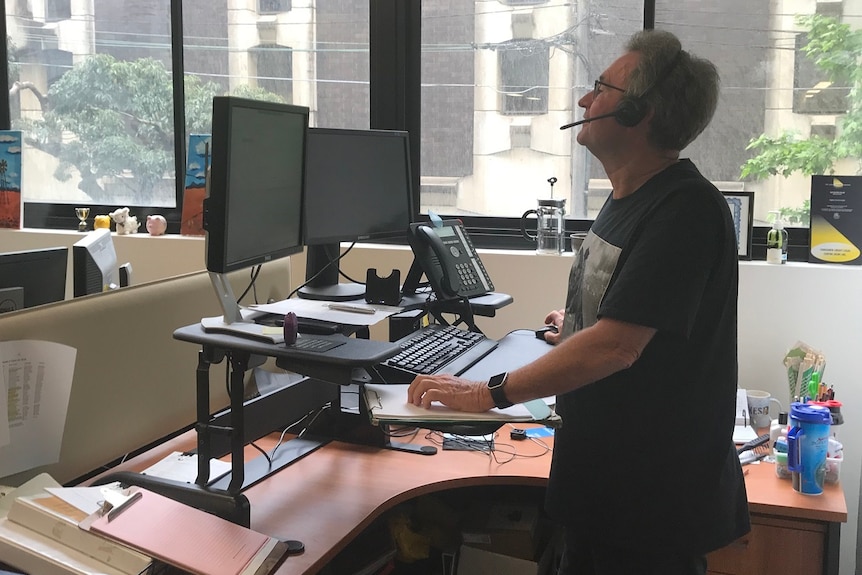 Man standing at raised work desk on the phone at a computer.