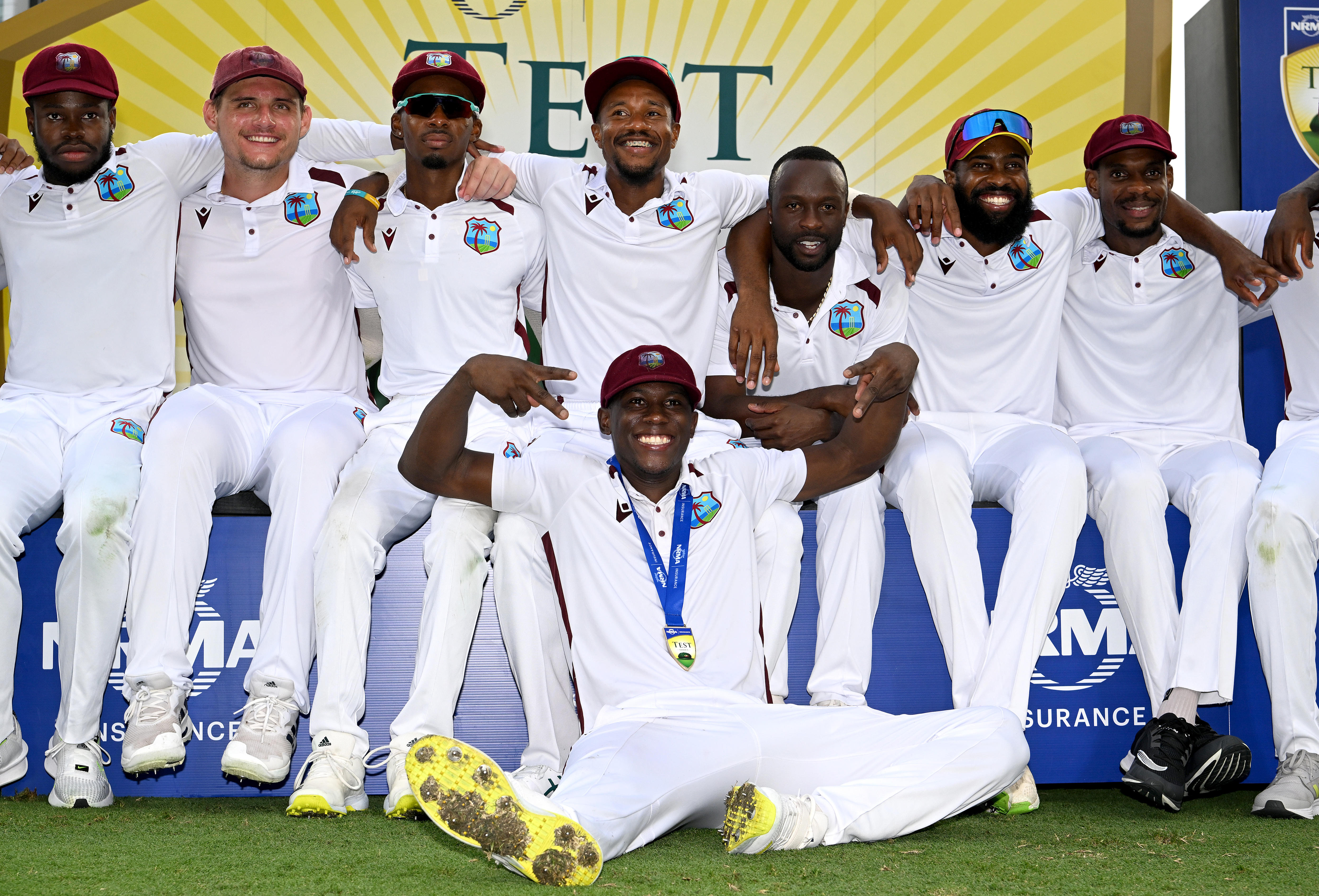 West Indies players sit together, arm in arm, with Shamar Joseph in front of them wearing a medal