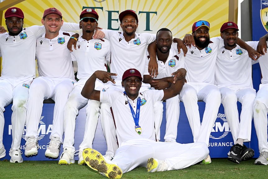West Indies players sit together, arm in arm, with Shamar Joseph in front of them wearing a medal
