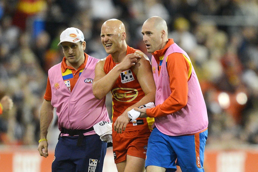 A grimacing AFL player holds his shoulder in pain as he is helped from the ground after an injury.