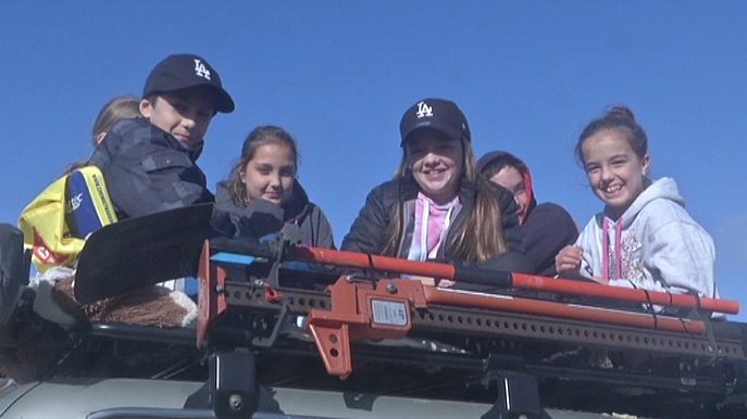 Six children sit on top of the roof racks of a four-wheel drive.