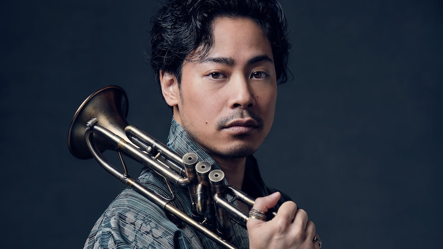 A tightly-cropped photo of trumpeter Jun Iida with his horn over his shoulder