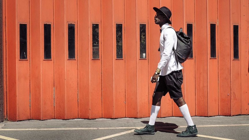 A man wearing black shorts, a long-sleeve white shirt and a black hat walks on a sunny street to depict wearing shorts at work.