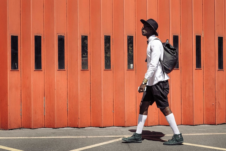 A man wearing black shorts, a long-sleeve white shirt and a black hat walks on a sunny street.