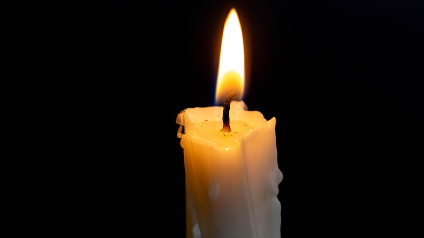 A white candle, burning, against a  black background