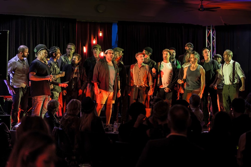 A group of men stand on a lit stage, singing