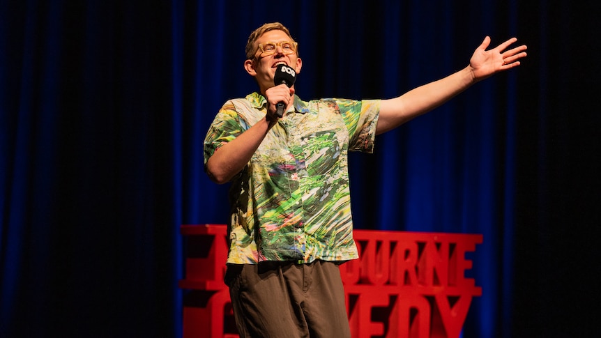 Chris Parker on stage with a red Comedy Festival sign and dark blue stage curtains in the background.