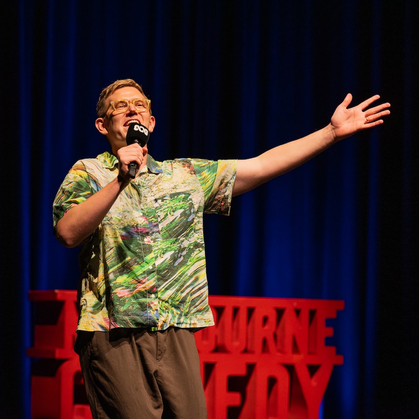 Chris Parker on stage with a red Comedy Festival sign and dark blue stage curtains in the background.