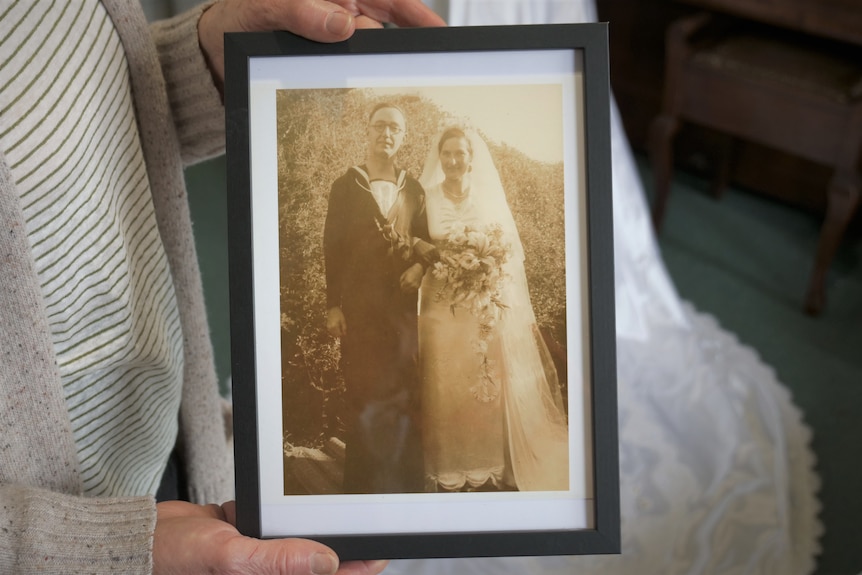 A pair of hands are shown holding a framed sepia photo of a bride and groom, the groom wearing naval uniform.