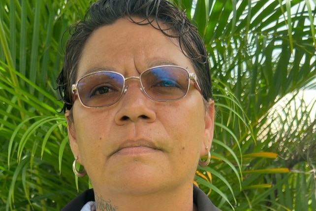 Una mujer indígena con gafas se para frente a las hojas de palma con una mirada decepcionada en su rostro