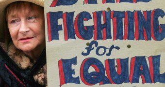 A woman holds a sign reading 'still fighting for equal pay'.