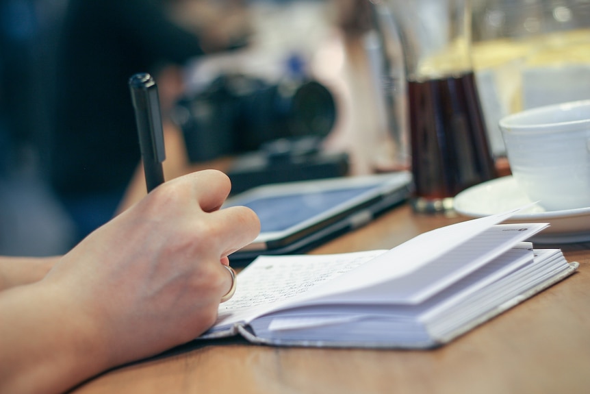 A hand writing in a paper journal next to a coffee