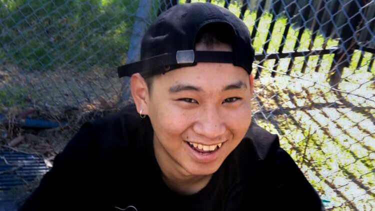 Dillon Wu, smiling, wearing a black backwards cap and black t-shirt and short, crouches in front of a fence