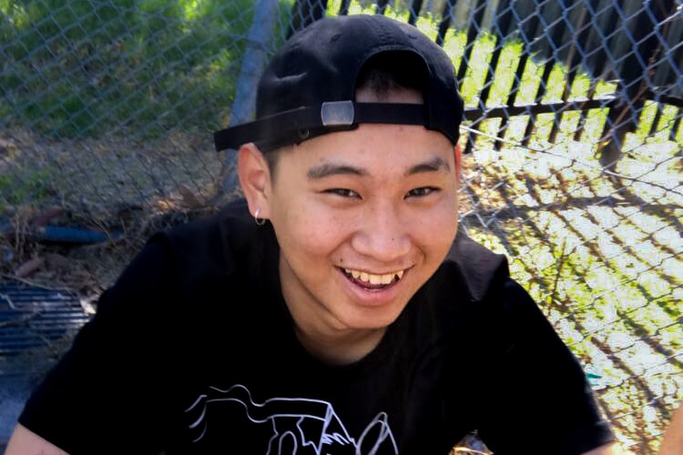 Dillon Wu, smiling, wearing a black backwards cap and black t-shirt and short, crouches in front of a fence