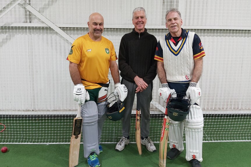 three men stand in front of cricket nets, all holding cricket bats