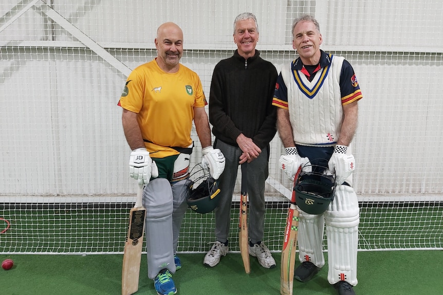 three men stand in front of cricket nets, all holding cricket bats