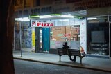 A person sitting on a bench at night looking at their phone outside a shop called Preston Pizza.
