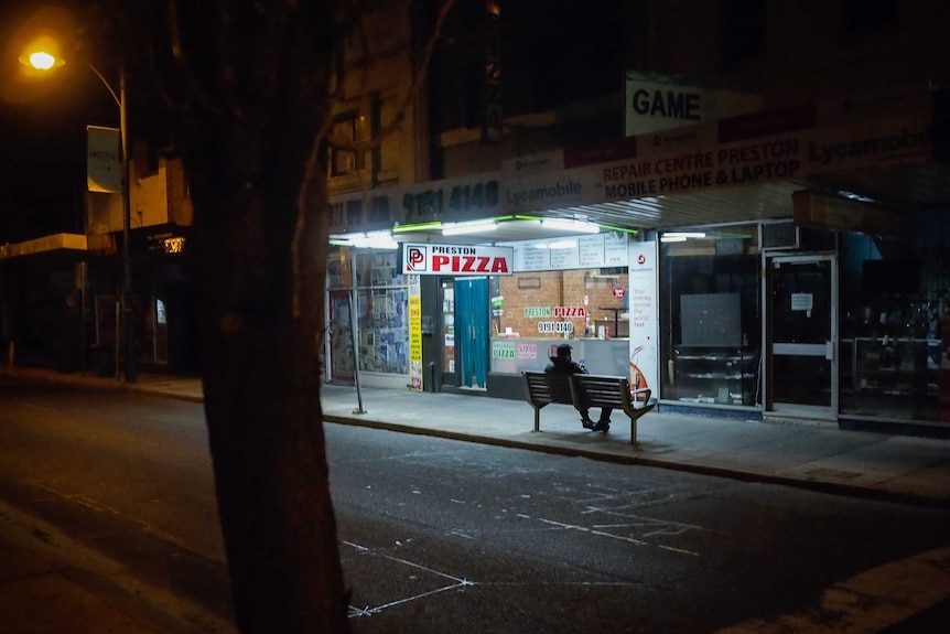 A person sitting on a bench at night looking at their phone outside a shop called Preston Pizza.