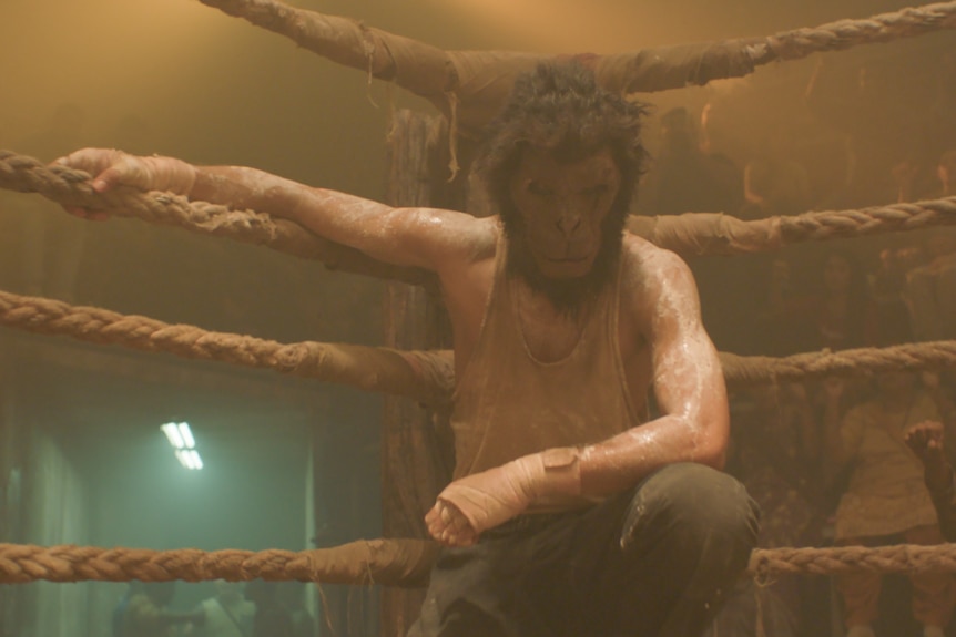 A film still of Dev Patel, wearing a gorilla mask. He is crouched in the corner of a wrestling ring, one arm on the ropes.