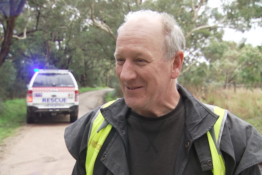 A man wearing a yellow high-vis vest over black clothing, with an SES vehicle behind him