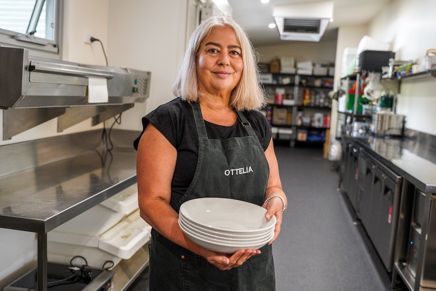 A woman wearing a black apron stands in a commercial kitchen carrying stack of plates.