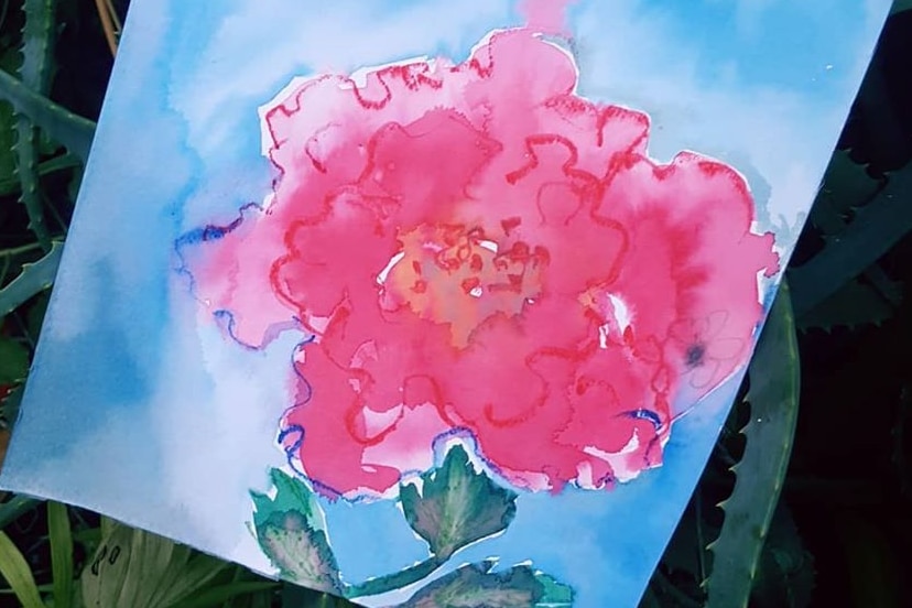 Against green plants sits a watercolour painting of a pinkish flower with green stem on a blue background.