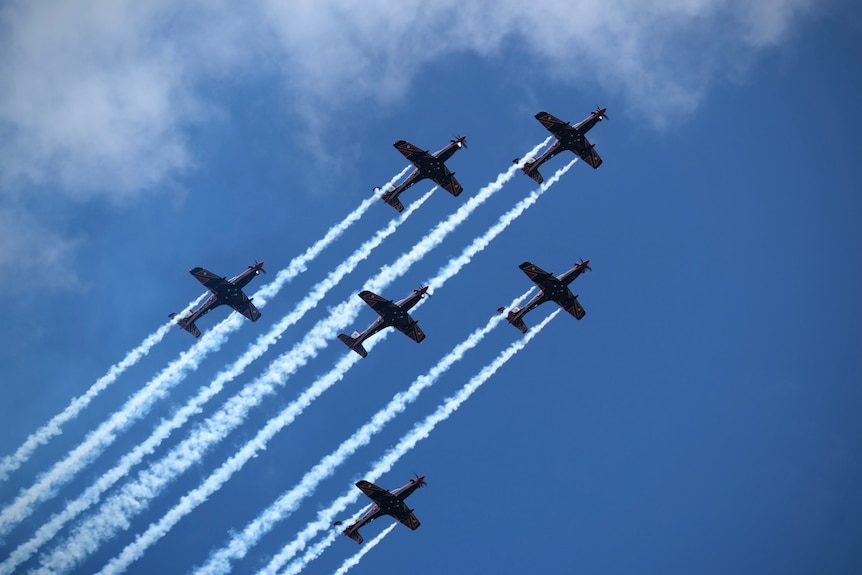 Six planes fly in formation against a blue sky.