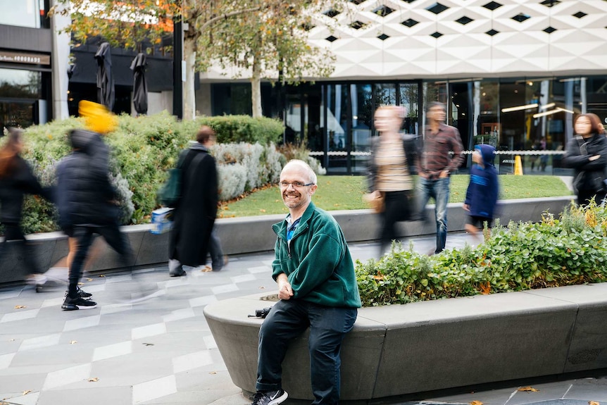 Short statured man Rob Paton sits next to a footpath, members of the public passing by behind him.