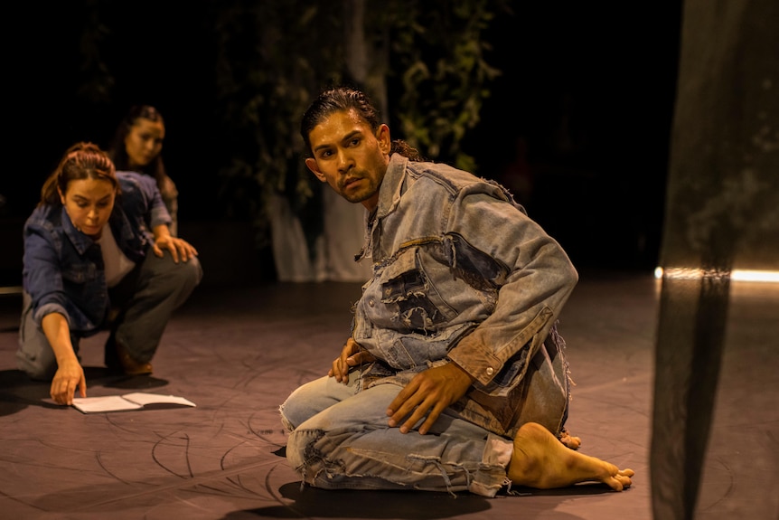 An Indigenous Australian man wearing all denim kneels on a stage. Two women in casual-wear are crouching in the background.