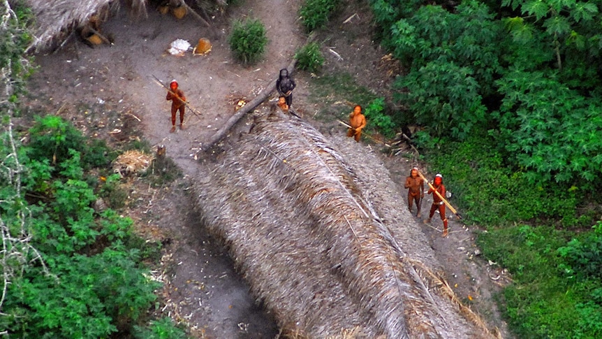 Five indigenous tribe members stand near a hut with spears in the Amazon