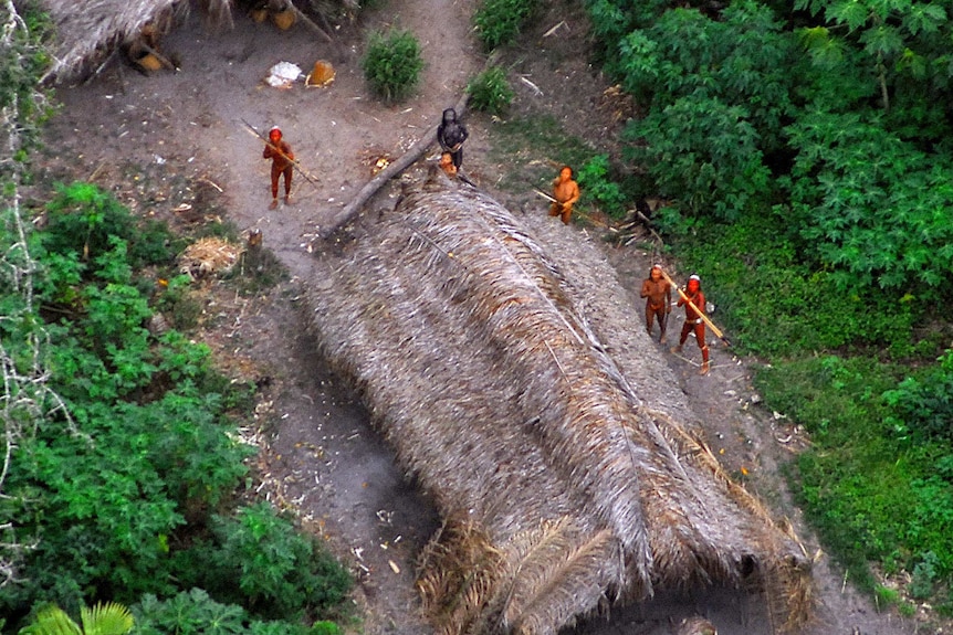 Five indigenous tribe members stand near a hut with spears in the Amazon