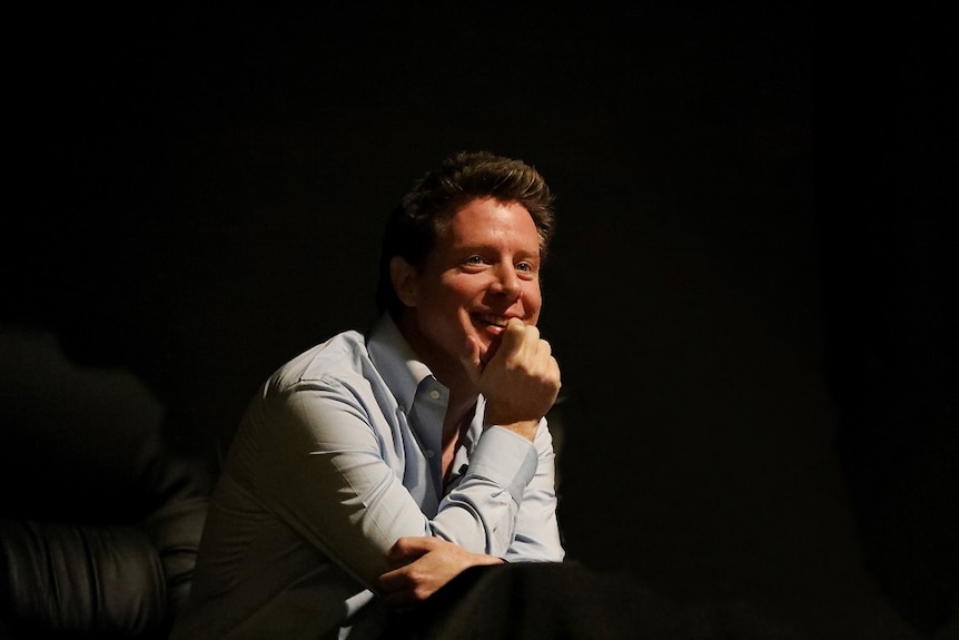 A young man, wearing a blue button-up shirt, smiles in a dark theatre